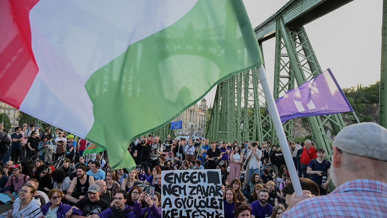 Un manifestante sostiene la bandera húngara mientras otros manifestantes se sientan en la calle junto al Puente de la Libertad durante una marcha en Budapest el 22 de abril de 2025, en respuesta a la aprobación por parte del parlamento húngaro de la reforma constitucional el 14 de abril de 2025. (Attila Kisbenedek / AFP vía Getty Images)