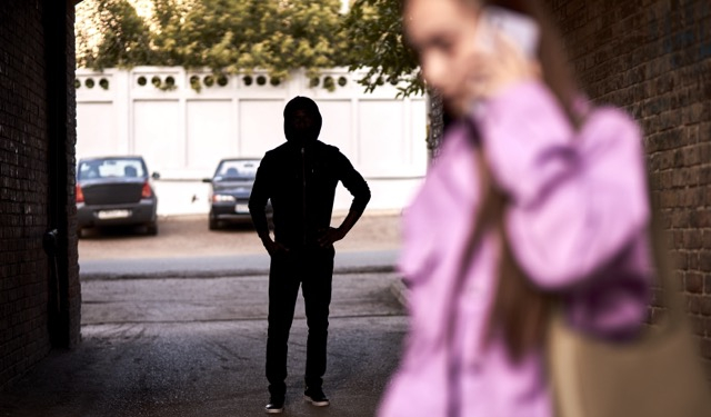 Criminal stalking a woman alone in dark street alley. Female is talking on phone. Unrecognizable man is hiding in the shadows. Woman Doesn't Suspecting That She Is Being Followed By Criminal Bandit